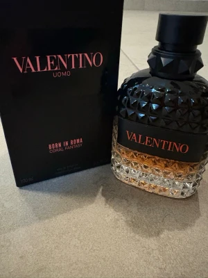 Valentino Uomo Born In Roma parfym - Valentino Uomo Born In Roma Coral Fantasy parfym i en lyxig glasflaska med nitar och svart lock. Flaskan har en snygg färgskiftning från klar till bärnstensfärgad och svart, med Valentino-loggan i rosa. Kommer med originalkartong.