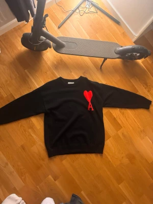 Svart stickad tröja från Ami - Svart stickad tröja från Ami Alexandre Mattiussi med röd hjärtlogga och bokstaven A broderad på bröstet. Tröjan har rund halsringning och långa ärmar. Perfekt för dig som gillar stilrena plagg med en twist.