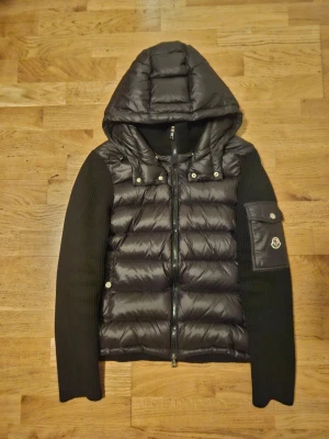 Svart Moncler hybridtröja med huva - Snygg svart hybridtröja från Moncler med vadderad front och rygg i blankt material samt stickade ärmar. Tröjan har en stor huva, dragkedja framtill och en ficka med Moncler-logga på ärmen. Perfekt för lager-på-lager och streetwear.