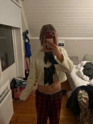 Vit paljettkofta med svarta rosetter - Säljer en kort, vit kofta täckt med paljetter och två stora svarta rosetter framtill. Koftan har lång ärm och rund halsringning, samt ett glansigt innerfoder. Perfekt för dig som vill sticka ut och addera lite extra glitter till din outfit.