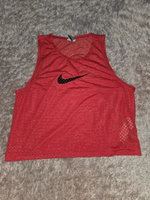 Röd meshtröja från Nike - Säljer ett rött mesh-linne från Nike med svart logga på bröstet. Perfekt för träning eller sport, tillverkat i lätt och ventilerande material. Modellen är ärmlös och har en avslappnad passform.