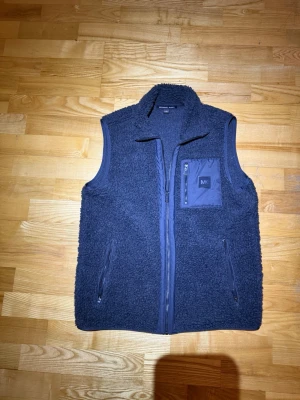 Svart  teddyväst från Michael Kors - Säljer en svart  teddyväst från Michael Kors med dragkedja framtill och tre fickor, varav en bröstficka med MK-logga. Västen är gjord i mjukt, fluffigt material och har en hög krage för extra värme. Perfekt för lager-på-lager och streetstyle.