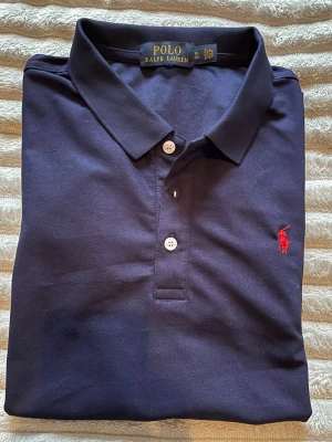 Mörkblå pikétröja från Ralph Lauren - Klassisk mörkblå pikétröja från Ralph Lauren med röd broderad logga på bröstet. Bra skick 