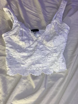 Vit spetsig linnetopp från Amisu - Superfin vit linnetopp från Amisu med spetsdetaljer och breda axelband. Toppen är croppad och har en romantisk känsla med v-ringning och blommigt spetsmönster. Perfekt att styla med jeans eller kjol för en trendig look.🩷