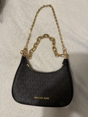 Svart handväska från Michael Kors - Snygg svart handväska från Michael Kors med guldfärgad kedja och logga framtill. Väskan har ett diskret mönster med MK-loggor och är tillverkad i slitstarkt syntetmaterial. Perfekt accessoar för att lyfta din outfit.