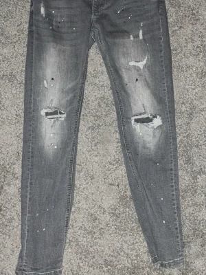 Grå skinny jeans med slitningar - Snygga grå skinny jeans med slitna knän och färgstänk för en trendig, avslappnad look. Jeansen har smal passform och är tillverkade i stretchigt bomullsmaterial. Perfekta för dig som gillar streetstyle och vill sticka ut lite extra.