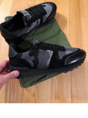 Valentino Garavani Rockrunner sneakers - Snygga Valentino Garavani Rockrunner sneakers i svart och grått med coolt kamouflagemönster. Skorna har snörning, platt sula och detaljer i mocka, skinn och textil. Perfekta för dig som vill ha en stilren men edgy look.