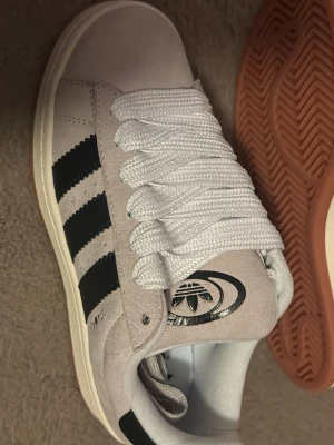 Adidas Campus sneakers beige och svart - Säljer ett par Adidas Campus sneakers i beige mocka med svarta ränder och vit sula. Skorna har breda vita skosnören och klassisk Adidas-logga på plösen. Yttersulan är i gummi med brun ton. Perfekta för dig som gillar retrostil.