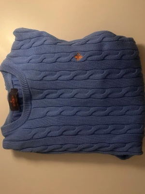 Blå kabelstickad tröja från Morris  - Snygg blå kabelstickad tröja från Morris med rund halsringning och diskret orange logga broderad på bröstet. Tröjan har klassiskt kabelmönster och ribbade muddar. Perfekt för en stilren och avslappnad look.