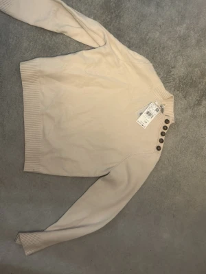 Beige stickad tröja med knappar - Snygg beige stickad tröja med ribbade muddar och hög hals. Tröjan har dekorativa bruna knappar längs axeln för en trendig touch. Perfekt att matcha med jeans eller kjol för en stilren look.