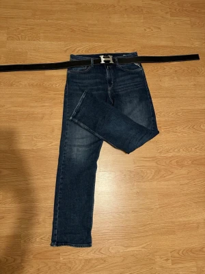 Mörkblå raka jeans med bälte - Snygga mörkblå jeans med raka ben och klassisk femficksdesign. Jeansen har en lätt tvättad look och kommer med ett svart bälte med silverfärgat H-spänne. Perfekta för en stilren och avslappnad outfit.