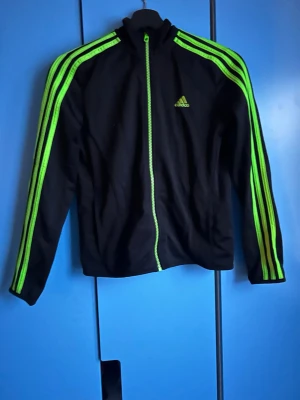 Svart Adidas träningsjacka med gröna detaljer - Svart träningsjacka från Adidas med neongröna ränder längs ärmarna och dragkedja framtill. Klassisk Adidas-logga på bröstet och hög krage. Perfekt för sport eller chill. Materialet är mjukt och följsamt.