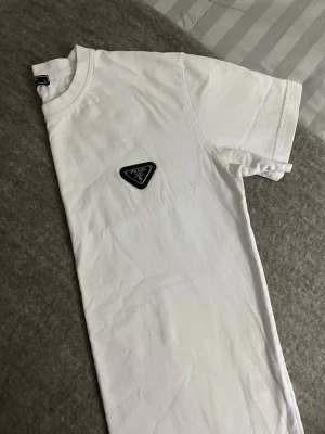 Vit t-shirt från Prada med logga - Vit t-shirt från Prada med klassisk rund hals och korta ärmar. På bröstet sitter en svart triangel-logga med Prada-tryck. T-shirten är i mjuk bomull och har en clean, stilren look som passar till allt.