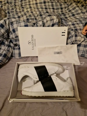 Valentino Opens - Snygga Valentino Garavani sneakers i vitt läder med svart band över ovandelen. Klassisk och stilren modell som passar till det mesta.  Skorna är använda, dem är smutsiga på sulan (se bild). Ovandelen är i bra skick utan större skador. Säljs med originalkartong och dustbag, vilket syns på bilderna.  Perfekta som vardagssneakers.