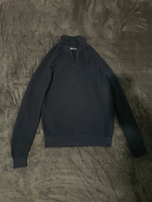 Mörkblå halvzip tröja - Mörkblå långärmad tröja med halv dragkedja och hög krage. Tröjan har ribbade muddar vid ärmslut och nederkant, vilket ger en snygg och bekväm passform. Perfekt för lager på lager och enkel att matcha med olika outfits.