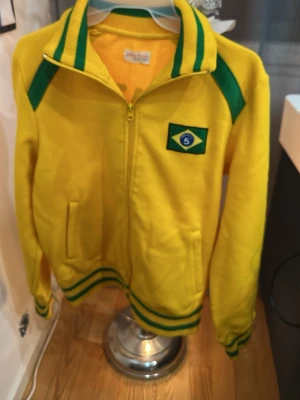 Gul Brasilien sportjacka med dragkedja - Gul sportjacka Jack & Jones  med gröna detaljer och dragkedja framtill. Jackan har Brasilien-flagga och nummer 5 på bröstet samt texten BRAZIL på ryggen. Ribbstickade muddar och krage med gröna ränder. Perfekt för fotbollsfans eller träning.