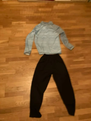 Nike träningsset med tröja och byxor - Nike träningsset med ljusblå långärmad tröja med halv dragkedja och vit logga på bröstet. Tillhörande svarta träningsbyxor med resår i midjan. Perfekt för löpning eller gym. Materialet känns lätt och andas bra.