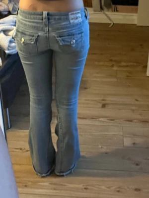 Ljusblå bootcut jeans med fickdetaljer - Säljer ett par ljusblå bootcut jeans med snygga fickdetaljer och knappar bak. Finns hål, men går säkert att sy/lappa ihop om man vill. Spilt färg men de syns knappt. 