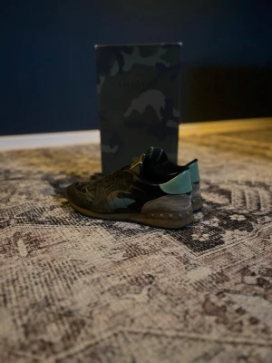 Valentino Rockrunner sneakers camouflage - Snygga Valentino Rockrunner sneakers med camouflage-mönster i svart, grön och grå. Skorna har detaljer i mocka och skinn, samt den ikoniska nitar på sulan. Perfekta för dig som vill ha en unik och trendig look.