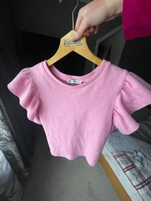Rosa topp med volangärmar från Zara - Supersöt rosa topp från Zara med stora volangärmar och rund halsringning. Croppad modell som ger en trendig look. Perfekt för dig som vill sticka ut lite extra! Materialet känns mjukt och bekvämt, troligen bomull eller en bomullsblandning.