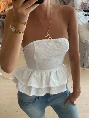 Vit axelbandslös peplum korsettopp - Säljer en vit axelbandslös korsettopp med peplumvolang och diskret blommönster. Toppen har en figurnära passform och är perfekt för att skapa en chic look till jeans eller kjol. Materialet känns som bomull eller polyester.