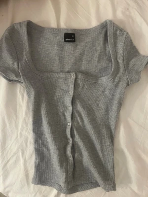 Grå ribbad topp från Gina Tricot - Grå ribbad topp med kort ärm och knappar framtill från Gina Tricot. Modellen är croppad med fyrkantig urringning och figurnära passform. Perfekt för en trendig och avslappnad look.