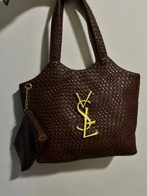 Brun shoppingväska från YSL i skinn - Snygg brun shoppingväska från YSL i flätat skinn med stort guldfärgat YSL-emblem framtill. Väskan har två rejäla handtag och en matchande liten clutch med guldkedja som kan fästas i väskan. Perfekt för dig som vill ha både stil och funktion.