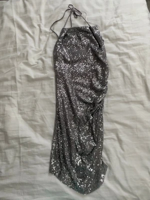 Glittrig silver halterneckklänning - Säljer en glittrig silverfärgad halterneckklänning med paljetter. Klänningen har smala knytband i nacken och en figurnära passform. Perfekt för dig som vill sticka ut på nästa fest eller event.