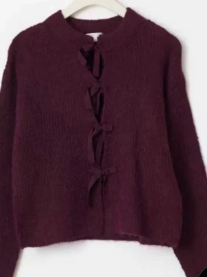 Vinröd stickad tröja med rosetter - Mysig vinröd stickad tröja med långa ärmar och unika rosettdetaljer framtill. Tröjan har en croppad passform och rund halsringning. Perfekt för dig som vill ha något extra i garderoben.