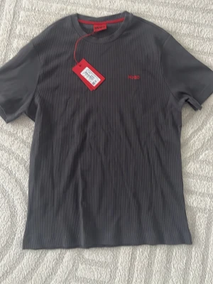 Mörkgrå ribbad t-shirt från Hugo - Snygg mörkgrå ribbad t-shirt från Hugo med diskret röd logga broderad på bröstet. T-shirten har korta ärmar och rund halsringning. Perfekt för dig som gillar stilrena och moderna plagg med en twist.