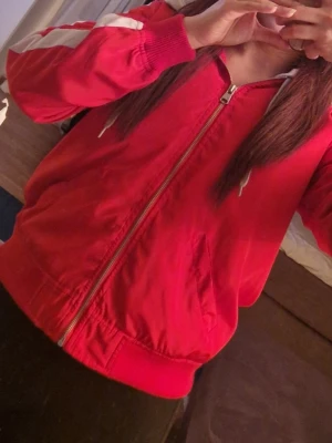 Röd bomberjacka med vita detaljer - Röd bomberjacka med vita ränder längs ärmarna och dragkedja framtill. Jackan har ribbade muddar vid ärmslut och midja, samt en klassisk bomberjacke-look. Perfekt för en sportig och avslappnad stil.