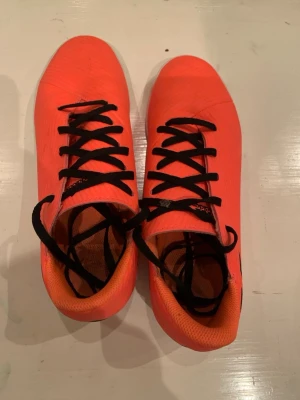 Orangea adidas fotbollsskor med snörning - Knappast använda som nya! Säljer ett par snygga orangea fotbollsskor från adidas med svarta ränder på sidan och svarta skosnören. Skorna har en låg profil och är tillverkade i syntetmaterial, perfekta för dig som vill sticka ut på planen.