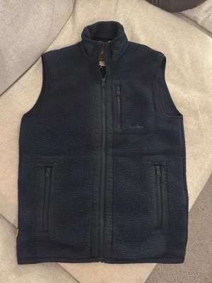 Svart fleeceväst från Timberland - Mysig svart fleeceväst från Timberland med hög krage och dragkedja framtill. Västen har tre praktiska fickor med dragkedja och detaljer i svart tyg. Perfekt att slänga på över en hoodie eller tröja för extra värme.