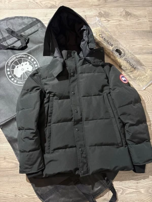 Canada Goose - Snygg svart dunjacka från Canada Goose med huva och klassisk logga på ärmen. Jackan är quiltad och har dragkedja samt knappar framtill. Medföljer avtagbar pälskant till huvan och originalpåse. Perfekt för kalla vinterdagar.