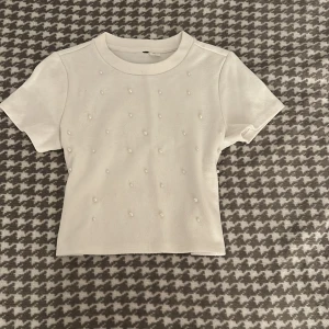 Vit croppad topp med pärlor - Tyvärr är den förliten. Du betalar för frakten och inga returneringar. Säljer en vit croppad topp med korta ärmar och rund hals. Toppen är dekorerad med små pärlor på framsidan för en söt och trendig look. Materialet är mjukt och stretchigt, perfekt för att styla med jeans eller kjol. Passformen är slim och toppen är enkel att matcha.