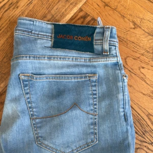 Ljusblå jeans från Jacob Cohën, strl 34 - Säljer ett par ljusblå jeans från Jacob Cohën med klassisk femficksdesign, snygga kontrastsömmar och logga på bakfickan. Jeansen har knappgylf och patch med märkesnamn i blått på linningen. Perfekta för en avslappnad och trendig look.