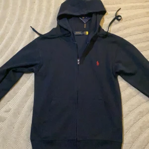 Mörkblå hoodie från Polo Ralph Lauren - Snygg mörkblå hoodie från Polo Ralph Lauren med röd broderad logga på bröstet. Tröjan har huva med snörning, dragkedja framtill och två fickor. Tillverkad i mjuk bomull, perfekt för chill dagar. Klassisk och stilren design som alltid funkar.
