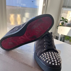 Christian Louboutin svarta sneakers med nitar - Svarta sneakers från Christian Louboutin i mocka med platt sula och rund tå. Tåpartiet är täckt av silvriga nitar för en edgy vibe. Skorna har snörning och den klassiska röda sulan. Perfekt för dig som gillar att sticka ut med din stil.(Sulans smutt går bort med ett blött papper) skorna passar även dig ner till 40,5