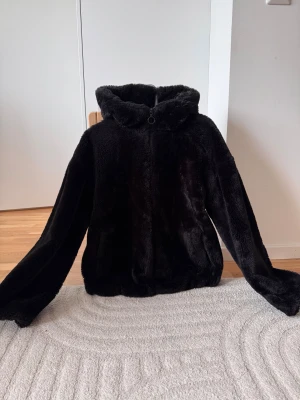 Svart teddyjacka från Primark - Mjuk och fluffig svart teddyjacka från Primark med huva och hel dragkedja framtill. Jackan har långärmade ärmar och är fodrad på insidan för extra värme. Perfekt för kalla dagar när du vill vara både chill och snygg.
