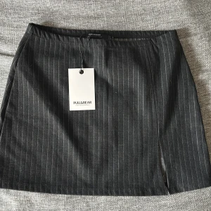 Svart randig minikjol Pull&Bear S - Svart minikjol från Pull&Bear med tunna vita ränder och en liten slits framtill. Kjolen har en rak passform och resår i midjan för extra komfort. Perfekt för dig som gillar en stilren och trendig look.