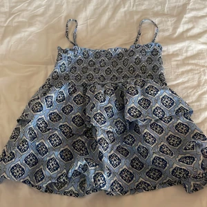 Blå mönstrad volang linne Gina Tricot -  Supersöt blått linne  från Gina Tricot med justerbara axelband som går att ta av och på och ett unikt mönster i mörkblått och vitt. Smockad överdel och volangig kjol ger en chill och trendig vibe. Perfekt för varma dagar och snygg att styla med sneakers eller sandaler. St 146/152 men passar xxs. Använd 1 gång