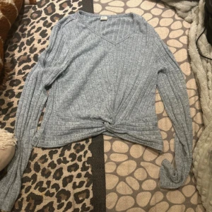 Grå ribbad långärmad tröja med knut - Snygg grå ribbad tröja med v-ringning och lång ärm. Tröjan har en cool knutdetalj framtill som ger en avslappnad vibe. Perfekt att matcha med jeans eller kjol för en chill look.