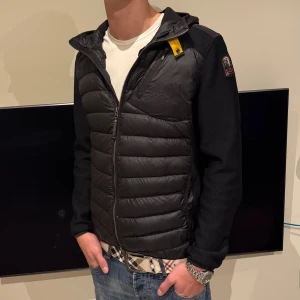  Parajumper cardigan  - Svart pufferjacka från Parajumpers med huva och coolt gult detaljspänne vid halsen. Jackan har quiltad framsida och rygg, meshade ärmar och axlar, samt dragkedja framtill. Märkespatch på vänster ärm och bröstficka med dragkedja. Perfekt för dig som gillar streetwear. Har storleken L men passar för M då modellen är M storlek. 