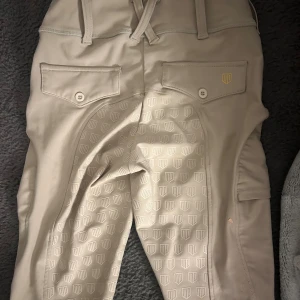 Beige ridbyxor Maya Delorez 2XS - Snygga beige ridbyxor från Maya Delorez i storlek 2XS. De har hög midja, praktiska fickor med knappar och ett mönstrat silikontryck för extra grepp. Materialet är stretchigt och bekvämt, perfekt för ridning. Diskret logga i guld på benet och bakfickan.