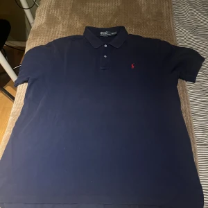 Marinblå pikétröja från Polo Ralph Lauren - Säljer en klassisk marinblå pikétröja från Polo Ralph Lauren i storlek XXL. Tröjan har krage, två knappar framtill och den ikoniska röda Polo-loggan broderad på bröstet. Tillverkad i mjuk bomull som är skön mot huden.