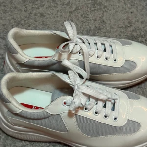 Vita American Cups sneakers   - Snygga sneakers i storlek 41 i vit och ljusgrå färg med rund tå och platt sula. Skorna har snörning och är tillverkade i en kombination av skinn och syntetmaterial med meshpaneler för extra komfort. Perfekt för dig som gillar stilrena och moderna sneakers.