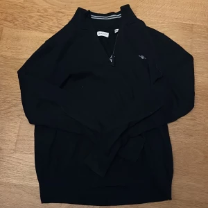 Svart half zip tröja från GANT - Svart långärmad tröja från GANT med half zip och broderad logga på bröstet. Tröjan har en ståkrage med diskret randig detalj på insidan och är tillverkad i mjukt bomullsmaterial. Perfekt för dig som gillar klassisk och stilren look. Köpt för 1350kr så detta priset jag satt är ganska rimligt om man tänker på hur ofta jag haft den på mig.