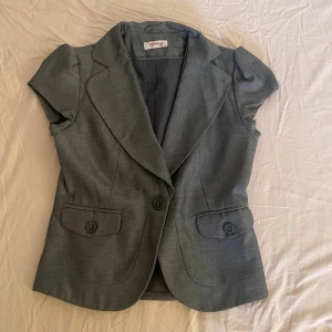 Grå kavajväst  - Snygg grå kavajväst med korta ärmar, breda slag och två framfickor med knappar. Västen har en figurnära passform. Strl XS