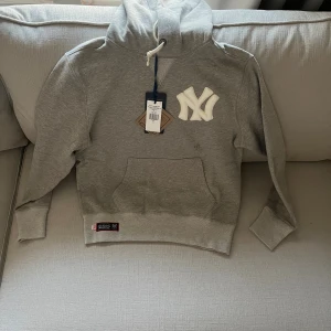 Grå hoodie Ralph Lauren Yankees S - Snygg grå hoodie från Ralph Lauren x MLB Yankees med broderad NY-logga fram och bak, samt stor RALPH LAUREN-text på ryggen. Tröjan har huva med snörning, känguruficka och ribbade muddar. Materialet är mjuk bomull och polyesterblandning. Slutsåld överallt! 