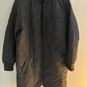 Svart quiltad lång bomberjacka - Säljer en svart lång bomberjacka med quiltat mönster och dragkedja framtill. Jackan har ribbade muddar vid ärmslut, fickor på sidorna och en bröstficka. Perfekt för dig som gillar stilren och enkel design med lite edge.